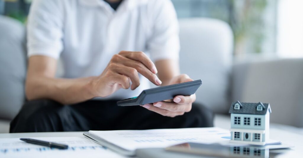 Person using a calculator near a small house model to plan home expenses, budgeting, and smart property investment decisions.